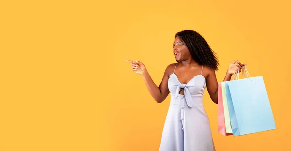 Excited young black woman with shopping bags pointing aside, offering big sale or unexpected promo on orange background