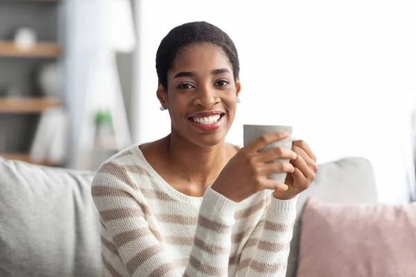 Ev Tatili. Gülümseyen siyah kadın koltukta oturuyor ve kahve içiyor. — Stok fotoğraf