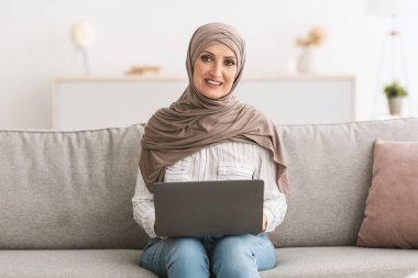 Üst düzey Müslüman kadın evde dizüstü bilgisayar kullanıyor, başörtüsü takıyor.