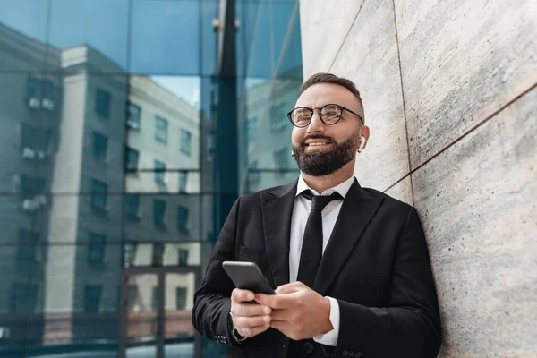 Happy office worker using smartphone and listening music in wireless earphones, standing near modern office building