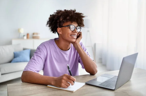 Trening online. Szczęśliwy czarny nastolatek uczy się zdalnie w domu, pisze w swoim notatniku, patrzy na ekran laptopa — Zdjęcie stockowe