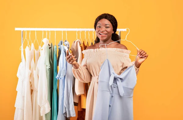 Gelukkig zwart vrouw met behulp van mobiele telefoon voor internet winkelen, plukken jurk in de buurt van kleding spoor, het kopen van de zomer garderobe — Stockfoto