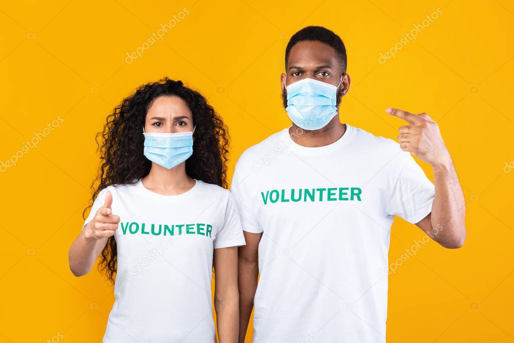Two Multiracial Volunteers Pointing At Their Face Masks, Yellow Background