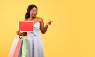 Elinde alışveriş poşetleri ve içi boş tabelayla turuncu arka plandaki boşluğu gösteren genç siyahi bir kadın.
