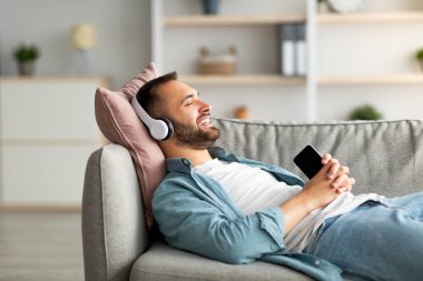 Havalı genç adam kanepede uzanıyor, kulaklık takıyor, cep telefonuyla müzik dinliyor, yeni uygulama kullanıyor, yer açıyor.