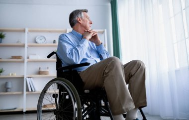 Tekerlekli sandalyedeki engelli, pencereden dışarı bakan, huzurevinde depresyonda olan yaşlı bir hastanın düşük açılı görüntüsü.