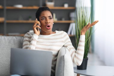 Endişeli siyahi kadın cep telefonuyla konuşuyor ve evde dizüstü bilgisayar kullanıyor.