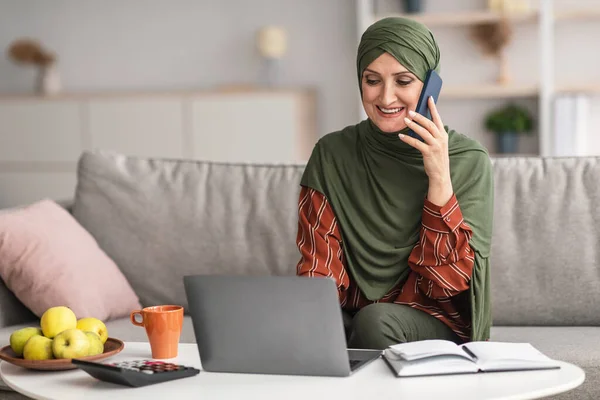 La señora islámica de mediana edad usando el ordenador portátil hablando por teléfono en interiores — Foto de Stock