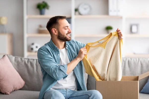 Щасливий чоловік клієнт відкриває картонну коробку, тримаючи свій новий светр, задоволений службою доставки вдома — стокове фото