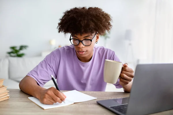Skupiony czarny nastolatek z filiżanką kawy robi notatki podczas wykładu online na laptopie w domu — Zdjęcie stockowe