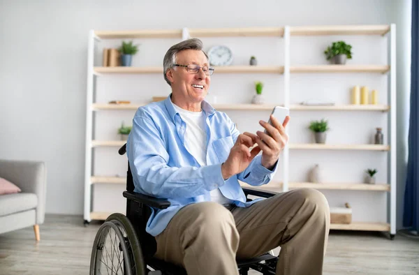 Happy disabled older man in wheelchair using cellphone, browsing web or watching movie at retirement home