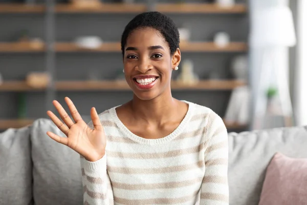 Friendly Black Woman Waving Hand At Camera, Sitting On Couch At Home