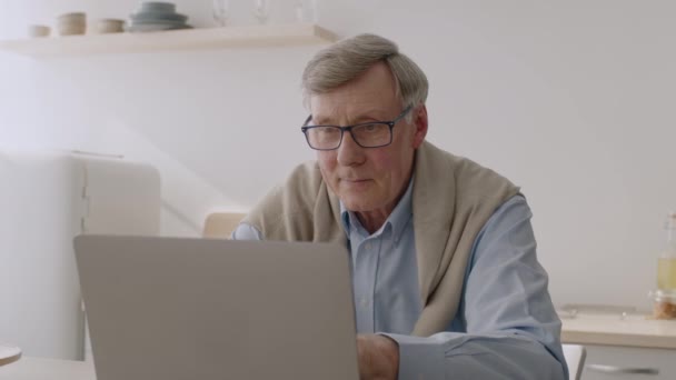 Freelance op pensioen. Vrolijke senior man in bril werkt op afstand in de keuken, lees project op laptop — Stockvideo