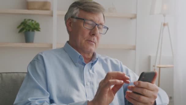 Homem sênior positivo vestindo óculos navegando no smartphone em casa e sorrindo, tiro de rastreamento, câmera lenta — Vídeo de Stock