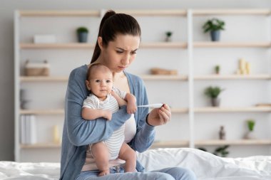 Endişeli genç anne yeni doğan bebeğinin sıcaklığını ölçüyor, termometreye bakıyor.