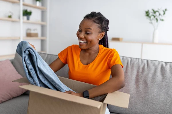 Auspacken der Kleidung. Porträt einer lächelnden schwarzen Dame, die Pappschachtel öffnet, neue Jeans herausnimmt und zu Hause auf dem Sofa sitzt — Stockfoto