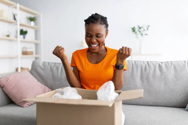 Izgatott fekete fiatal nő kibontása karton csomag a nappaliban, hogy IGEN gesztus, szereti a megrendelt termék — Stock Fotó
