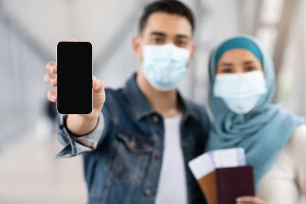 Pareja musulmana en máscaras médicas que demuestran Smartphone con pantalla negra en el aeropuerto —  Fotos de Stock
