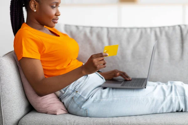 Happy black lady using laptop and credit card, sitting on couch, ordering products in web store. Online shopping concept