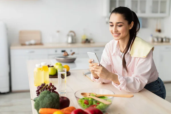 Porträt einer jungen Hausfrau, die ihr Handy an den Küchentisch lehnt — Stockfoto