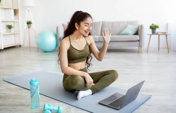 Jonge Indiase vrouw in sportkleding zittend op sportmat, begroet haar personal trainer online op laptop pc, binnen — Stockfoto