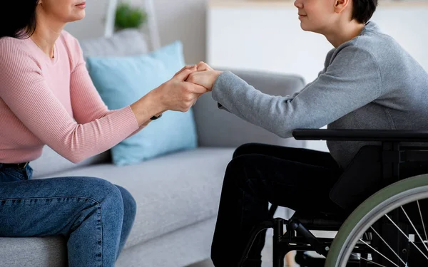 Інвалідний хлопчик-підліток у інвалідному візку отримує підтримку від матері або доглядальниці, тримаючи її за руки вдома, крупним планом — стокове фото