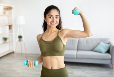 Gülümseyen Hintli kadın portresi ev içi eğitim yapıyor, dambıllarla egzersiz yapıyor, evde pazıları güçlendiriyor.