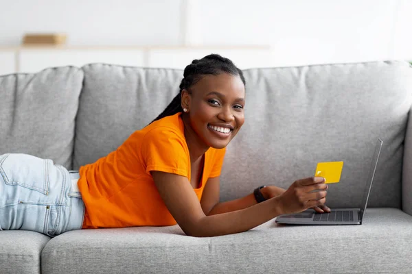 Czarna dama dokonująca płatności online, dokonująca zakupów w internecie, korzystająca z usług banku internetowego, leżąca na kanapie z laptopem i kartą kredytową — Zdjęcie stockowe