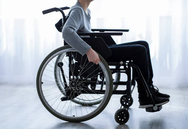Disability in teenagers concept. Side view of unrecognizable impaired teen boy sitting in wheelchair at home, closeup