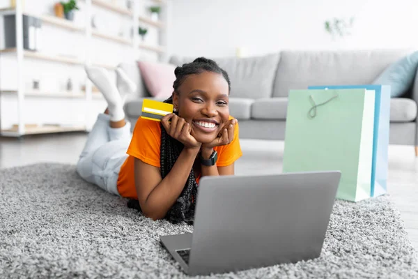 Glad ung svart kvinna som innehar kreditkort, med hjälp av bärbar dator, välja och köpa varor på webben, ligger på golvet inomhus — Stockfoto