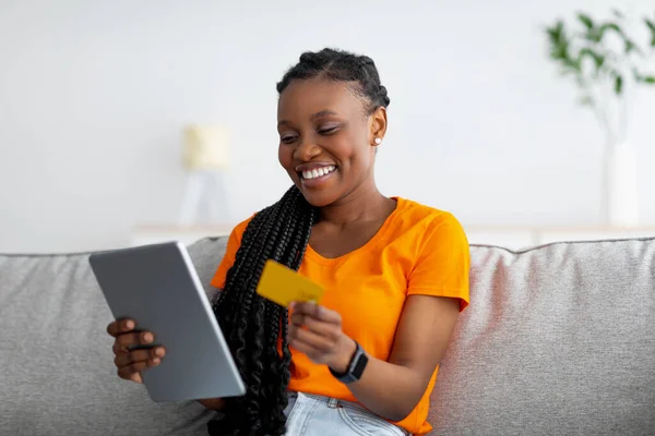 Donna afroamericana in possesso di carta di credito e utilizzando tablet pc, seduto sul divano a casa, shopping nel negozio online — Foto Stock