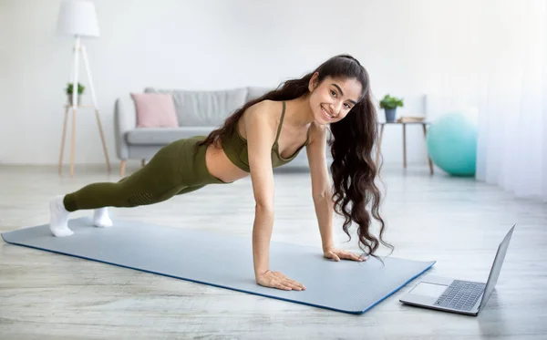 Glimlachende jonge Indiase dame doet plank, versterking van haar armen en lichaam in de voorkant van laptop thuis — Stockfoto