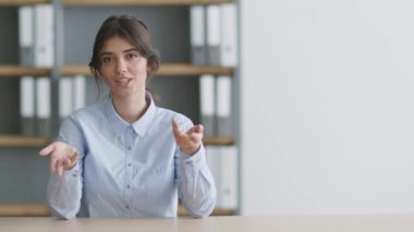 Genç profesyonel kadın konuşmacı kameraya konuşuyor, boşluğu gösteriyor ve işaret ediyor, ofis içinde oturuyor.