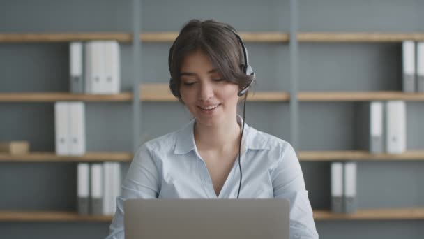 Koncepcja telepracy. Młody operator infolinii noszący słuchawki konsultingowe klienta zdalnie, mówiący i pracujący na laptopie — Wideo stockowe