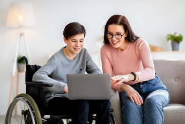 Evde eğitim kavramı. Tekerlekli sandalyedeki engelli çocuk annesiyle birlikte evde yeni online materyaller okuyor.