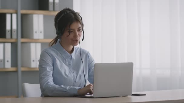 Nettbasert telefonsentral. Positiv ung kvinnelig operatør med hodetelefoner som snakker med kunden, som skriver på en bærbar datamaskin på kontoret – stockvideo