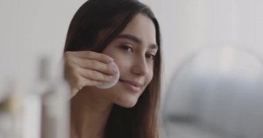Makyaj çıkarma ve cilt nemlendirici. Doğulu güzel bir kadın pamuklu bezle yüzünü temizliyor, aynaya bakıyor.