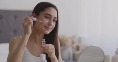 Yüzüne serum süren, aynaya bakan ve gülümseyen, spa ameliyatlarından zevk alan genç bir doğulu kadın.