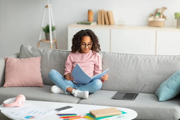 Концепція навчання та виховання. Усміхнена афроамериканська дівчина читає і робить домашнє завдання, сидячи на дивані з ноутбуком — стокове фото