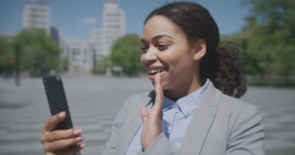 Comunicación a distancia. Alegre afroamericana dama vídeo chat con los socios a través de teléfono inteligente, de pie al aire libre — Vídeos de Stock