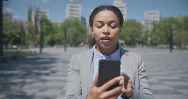Всегда на связи. Молодая занятая африканская американская деловая женщина в видеочате с деловыми партнерами через смартфон на открытом воздухе — стоковое видео