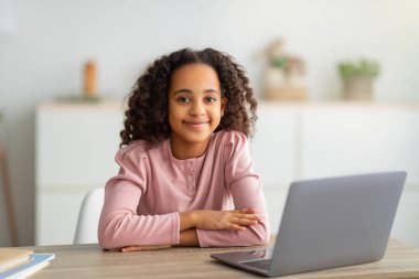 E-öğrenme ve bilgi. Masada oturan, dizüstü bilgisayar kullanan ve kameraya gülümseyen neşeli siyahi kız.