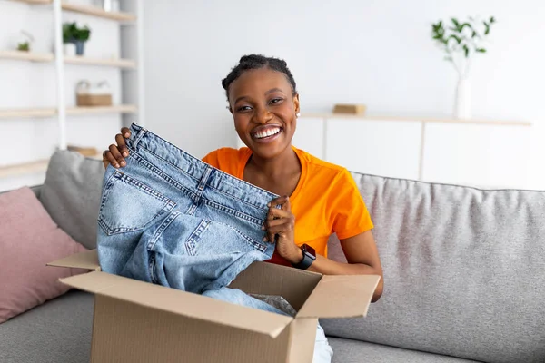 Glückliche schwarze Frau hält Jeans in der Hand, packt Pappverpackung aus, nimmt bestelltes Produkt in Empfang, ist mit ihrem Kauf zufrieden — Stockfoto