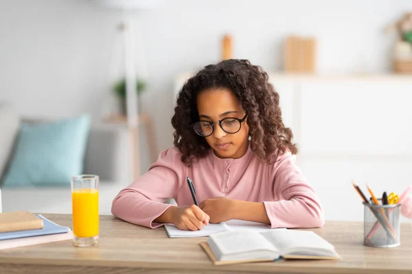 Nettes afrikanisch-amerikanisches Schulmädchen mit Brille, das in Notizblock schreibt, Hausaufgaben macht oder Essay schreibt, zu Hause lernt — Stockfoto