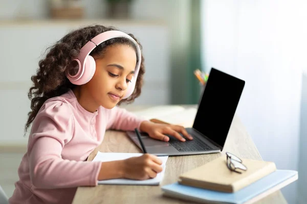 Adolescente negra con auriculares sentada en la mesa, usando un portátil con pantalla en blanco y escribiendo en su cuaderno — Foto de Stock