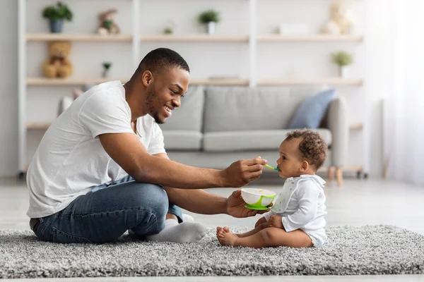 Licença de paternidade. Feliz pai preto alimentando bebê infantil de colher em casa — Fotografia de Stock