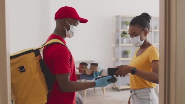 Black delivery man in protective mask giving coffee order to customer, lady paying for drink with mobile banking app — Stock Video