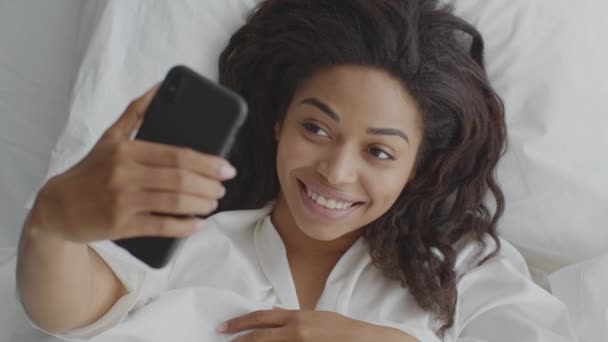 Genç Afro-Amerikalı bir bayan telefonda selfie çekiyor, sabahları yatakta uzanıyor, sosyal medya bağımlılığı, üst tabaka. — Stok video