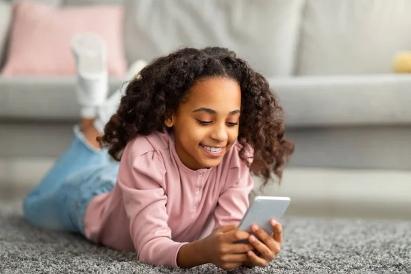 Digitale inboorlingen. Positief Afrikaans Amerikaans meisje met behulp van haar smartphone, sms 'en in sociale media, liggend op vloerkleed — Stockfoto