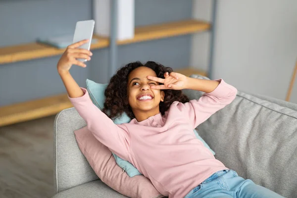 Legal afro-americano adolescente menina tomando selfie para a rede social, mostrando v sinal gesto, deitado no sofá em casa — Fotografia de Stock